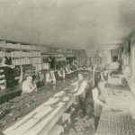 Workers are busy on the assembly line of the cutting room floor. The left side wall has shelving with white boxes stocked on it.