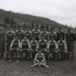 3 coaches and 6 players stand behind 6 seated players, while one sits cross legged at the front, with two baseball bats criscrossed on his lap making an 'X'.