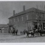 Large two story brick building with 5 windows on top level, and two chimney stacks. In the forefront, there are two carriages being pulled by horses down a dirt road as some young people stand outside the store.
