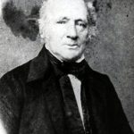 Black and white portrait of George Rolph as faces the camera. He is clean shaven with white hair and a black cloak.