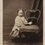 A young child stand beside a chair holding a book.