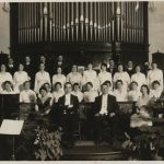 group of 32 men and women arranged in four rows, some sitting and some standing. Organ pipes are in the background while a piano sits in the foreground.