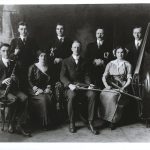 8 people with instruments--4 men stand behind 2 men and 2 women seated. There are 2 clarinets, 2 trumpets, 2 violins and 1 double-bass.