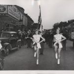 Two girls twirl batons as they dance in a parade past the theatre entrance. It is on the left side and now named the Majestic Theatre.