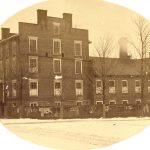 sepia postcard of a brick building. The left side shows a staircase and entrance to a three story building with a two story section attached at the back.