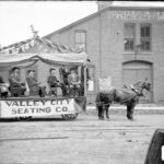 Eight men sitting on pews atop a decorated wagon for the Valley City Seating Company. They are being pulled by two horses in the parade.