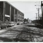 In the foreground there are rail tracks and cars parked along the street. To the left there is a large brick building with a steel fence around the property and an electrical structure with wires that cross over the road to the streetlight.