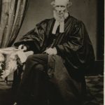 Older man sitting with his left leg over his right. His hair is balding and has a beard, wearing a Geneva gown over a cassock with white preaching tabs.