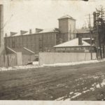 with the cotton mill complex in the background, there is a small building, titled Chapman Engine and MFG Co. There is a wooden fence around it and along the road.