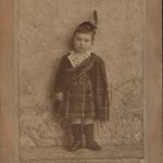 sepia photograph of James at age 2 wearing a traditional Scottish outfit. He wears a cap on his head with a feather and tartan kilt.