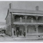four men and a dog stand outside the entrance while four women stand on the second floor veranda. There is a man and a horse standing at the back of the building, down the laneway.