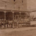 three horses pull a wagon with 5 people sitting on it. Two men stand in front of the horses. Men line along the lower veranda and entrance of the hotel as one man stands on the top veranda.