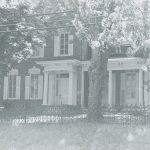 black and white photo of an Italianate Revival style house. There are white pillars at the entry and a iron fence around the property.