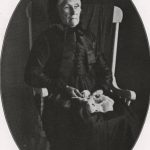 black and white portrait of Jane Giles in her senior years. She sits in a wooden rocking chair, wearing a black dress and hat, holding what appears to be knitting yarn and needles.