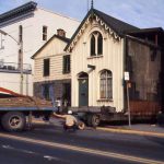 A pale yellow building on a flatbed trailer being towed onto King Street. A small crowd gathers to the right.